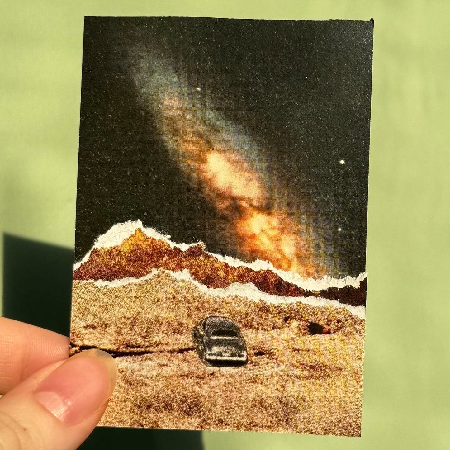 Hand holding a small collage of a car on a road with a celestial event in the sky.